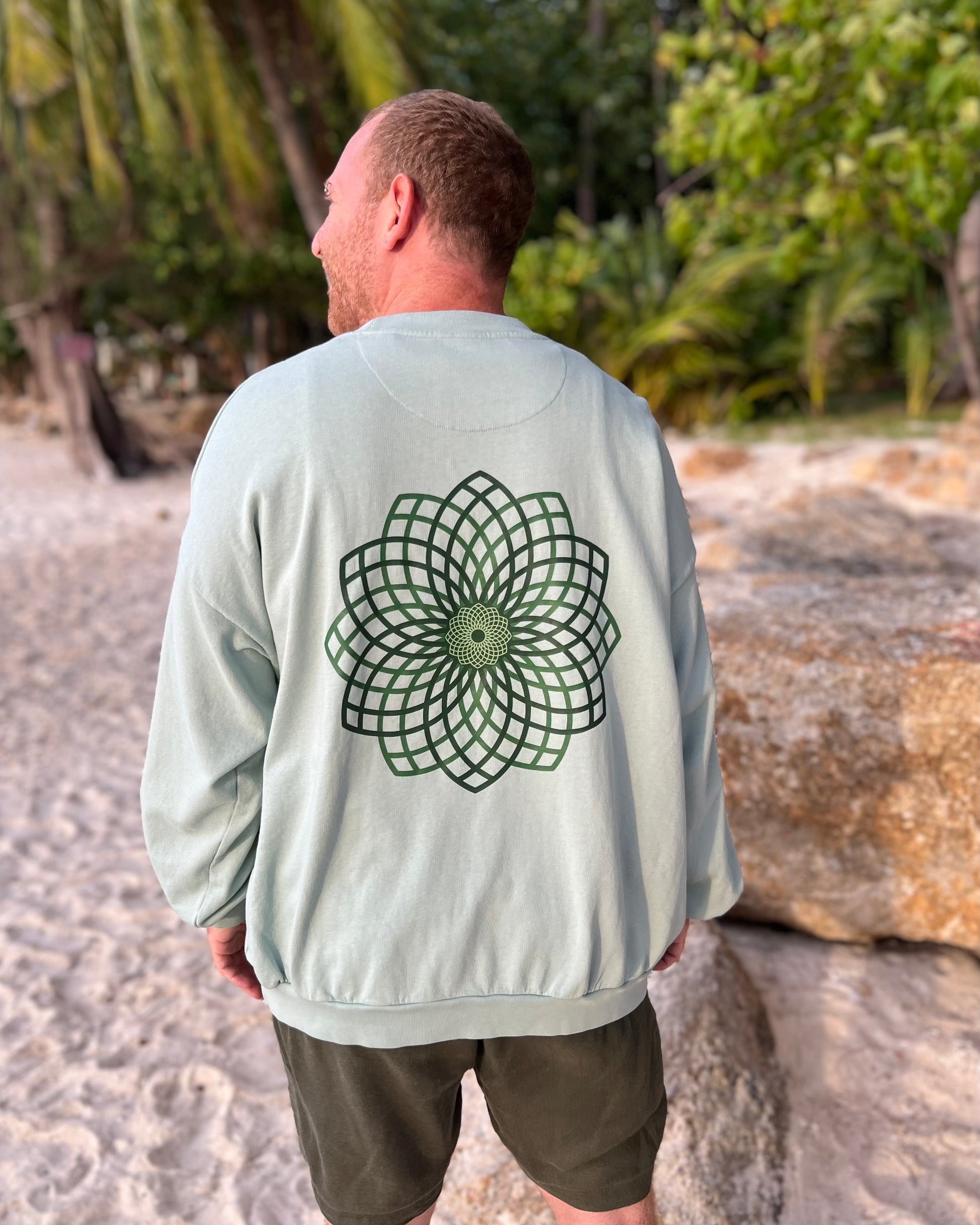 Man wearing a light green sweatshirt with a geometric flower design on the back, standing on a beach.