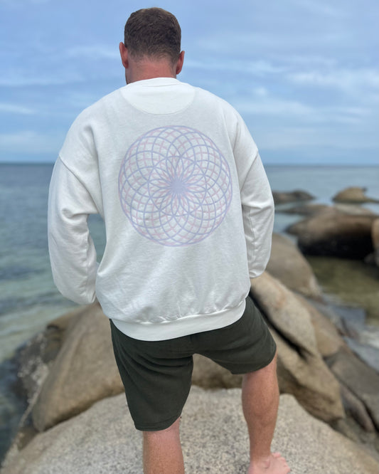 Person wearing a white sweatshirt with a geometric design on the back, standing on rocks by the ocean.
