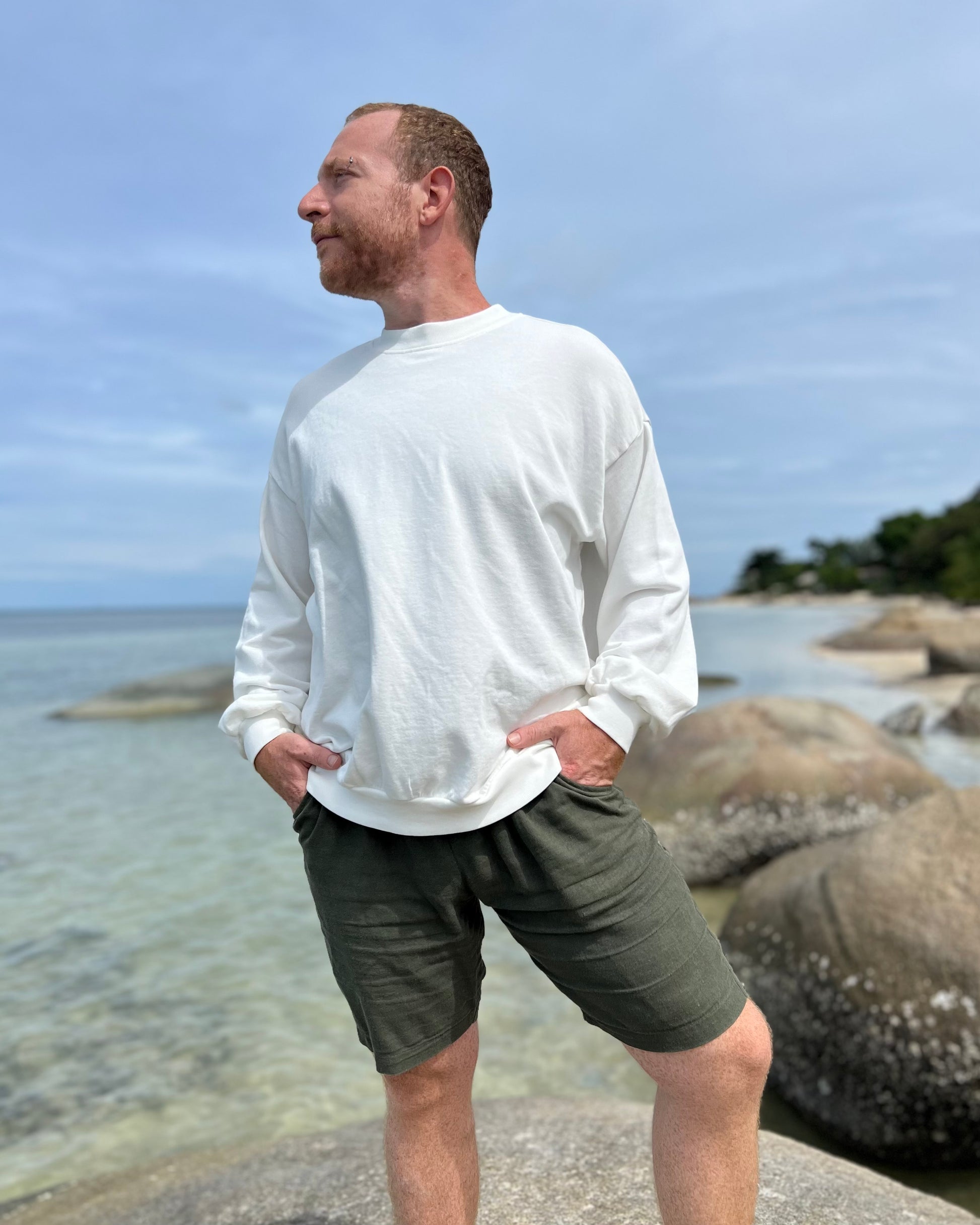 Man wearing a white long-sleeve oversized sweatshirt shirt standing on a rocky beach.