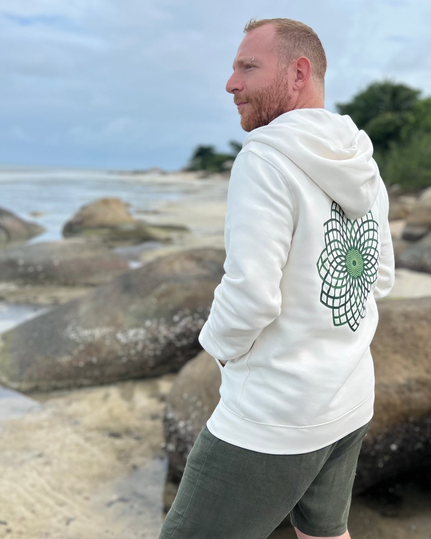 Man wearing a white hoodie with a green geometric design on a rocky beach.