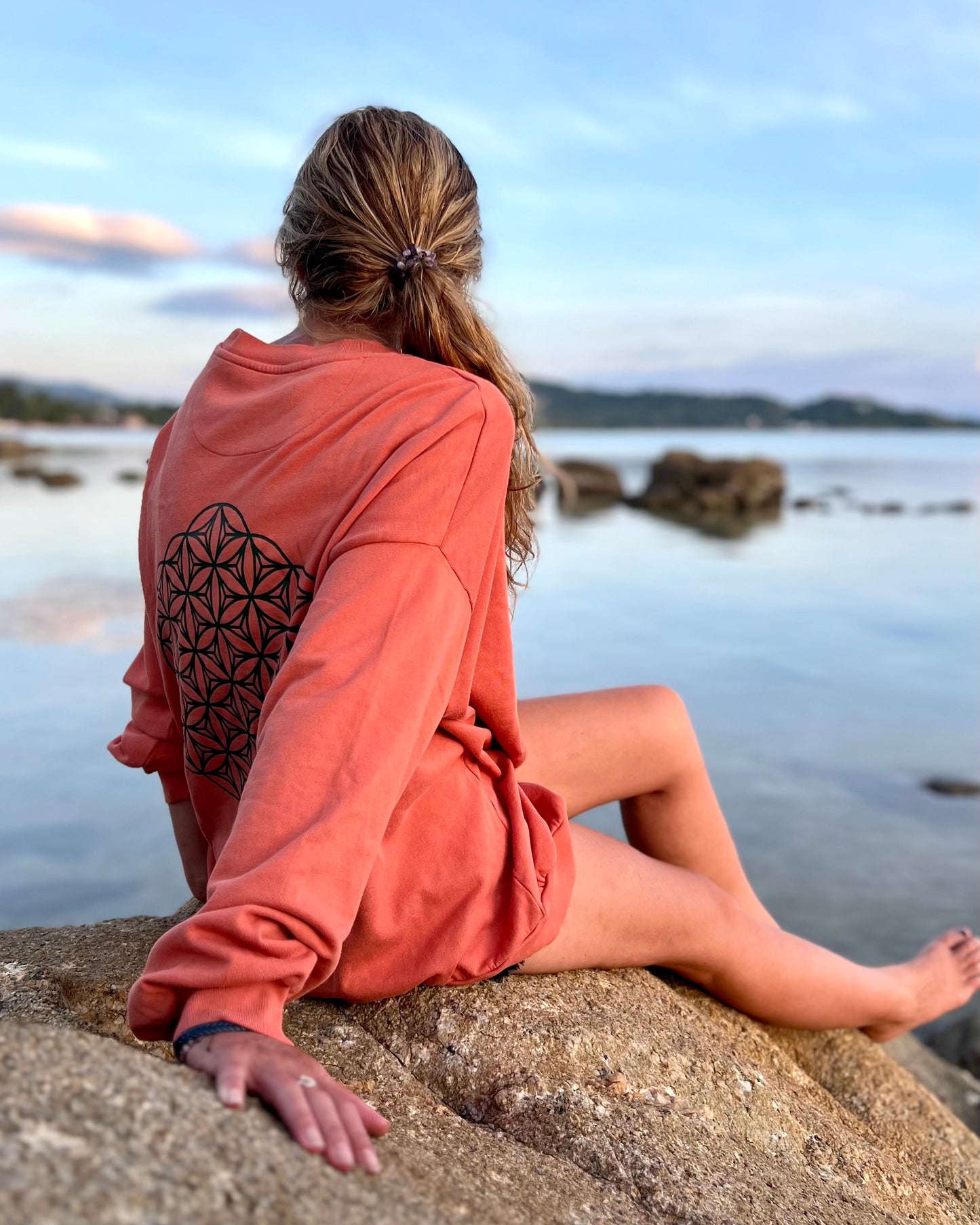 Person sitting on a rock by the water wearing an orange hoodie with a geometric design.