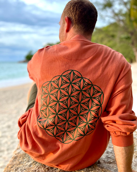 Person wearing an orange sweatshirt with a geometric design sitting on a beach.