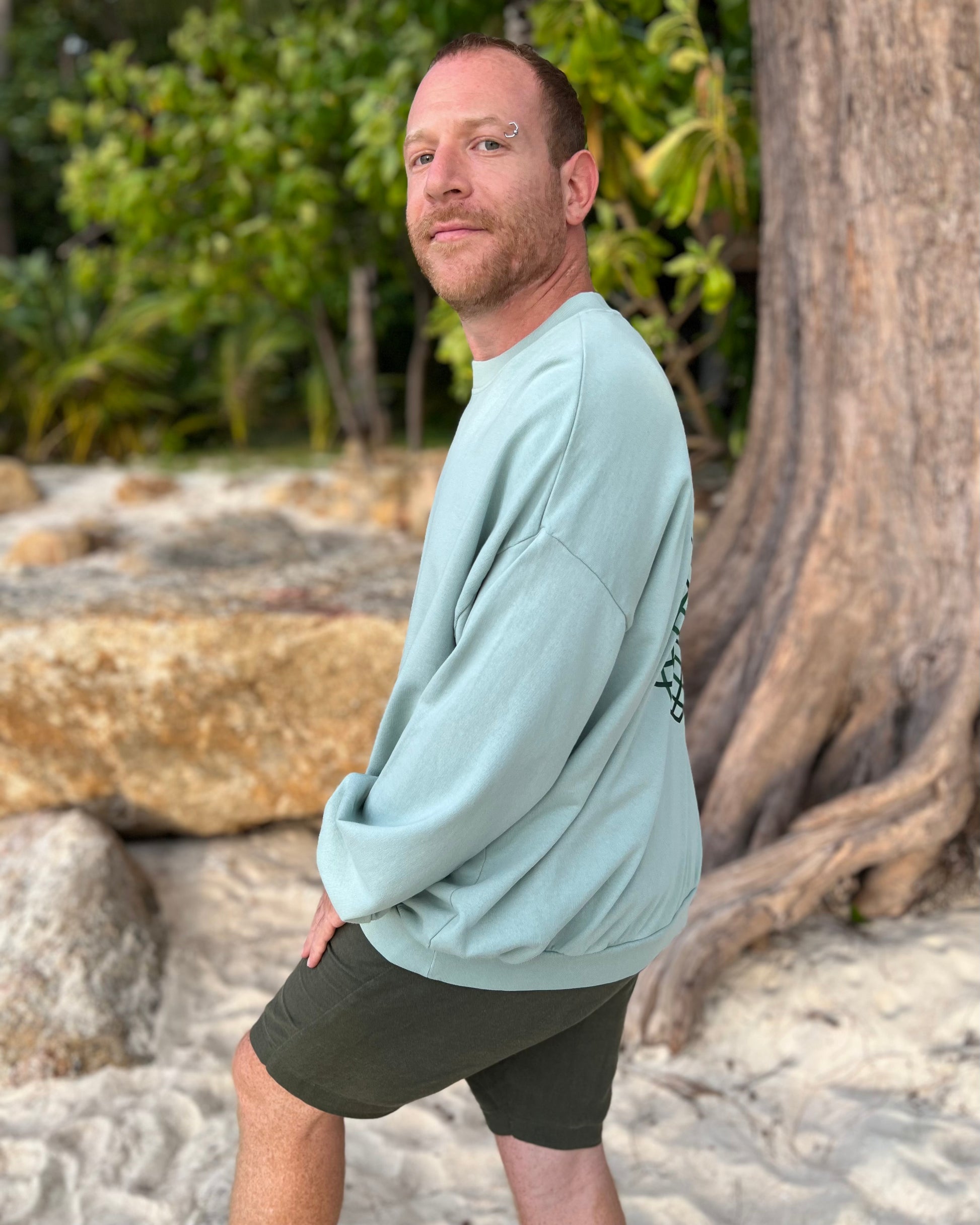 Man wearing a light blue sweatshirt and dark shorts standing on a rocky beach with trees in the background.