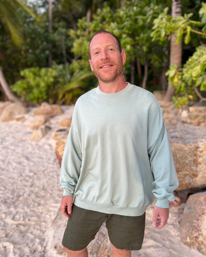 Man wearing a light blue sweatshirt and green shorts standing on a rocky beach with trees in the background.