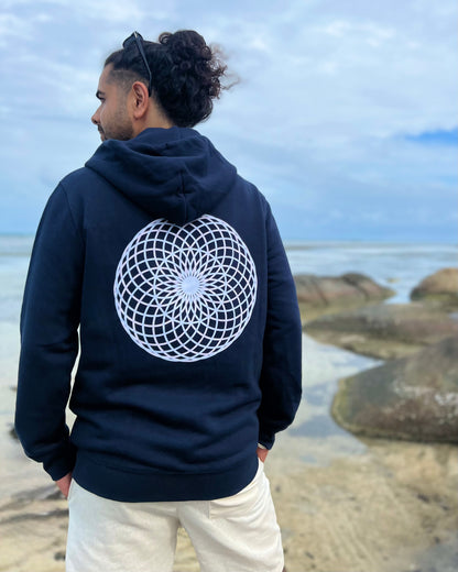 Person wearing a navy hoodie with a white geometric design on the back, standing by a coastal area.