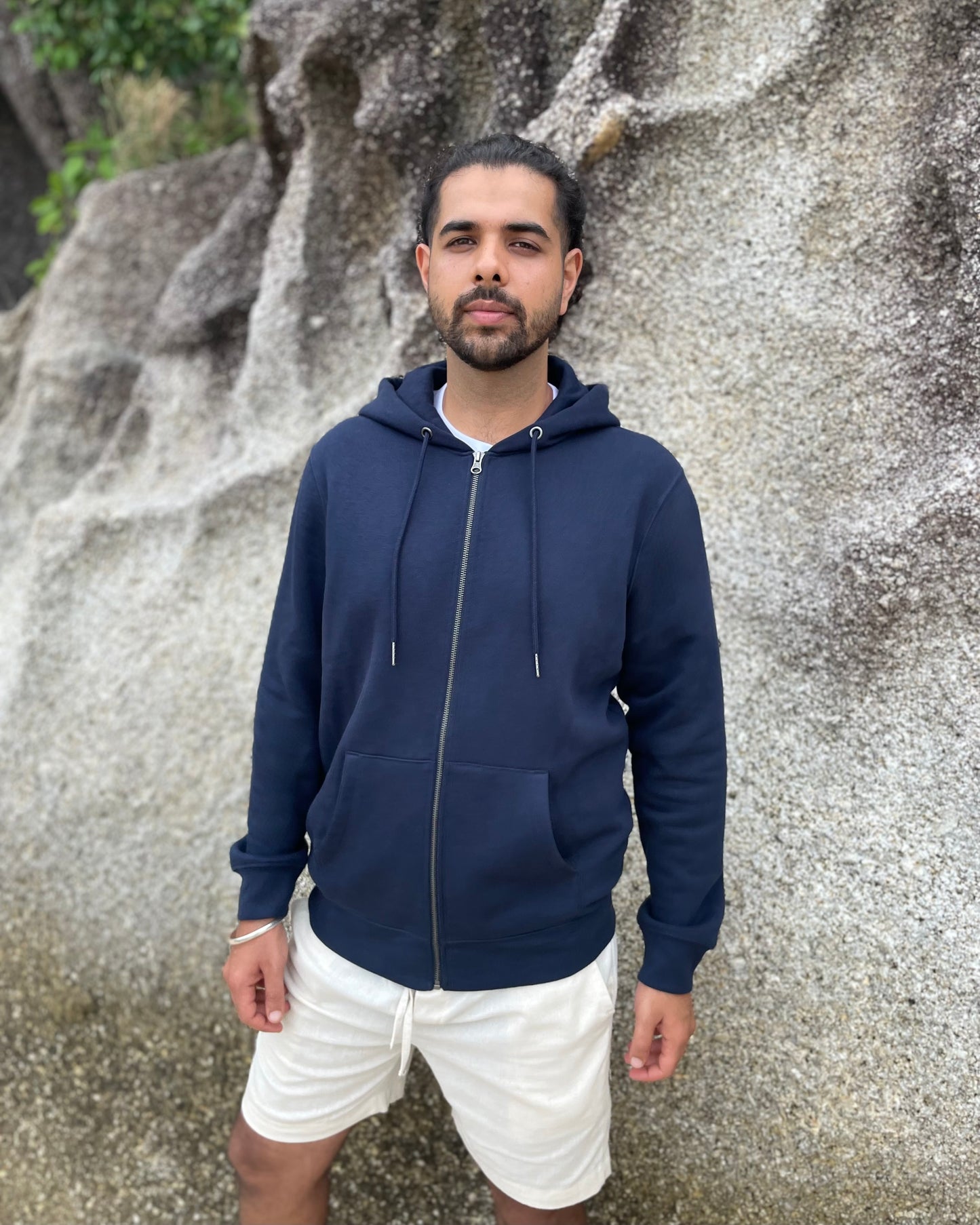 Man wearing a navy blue hoodie and white shorts standing against a rocky background