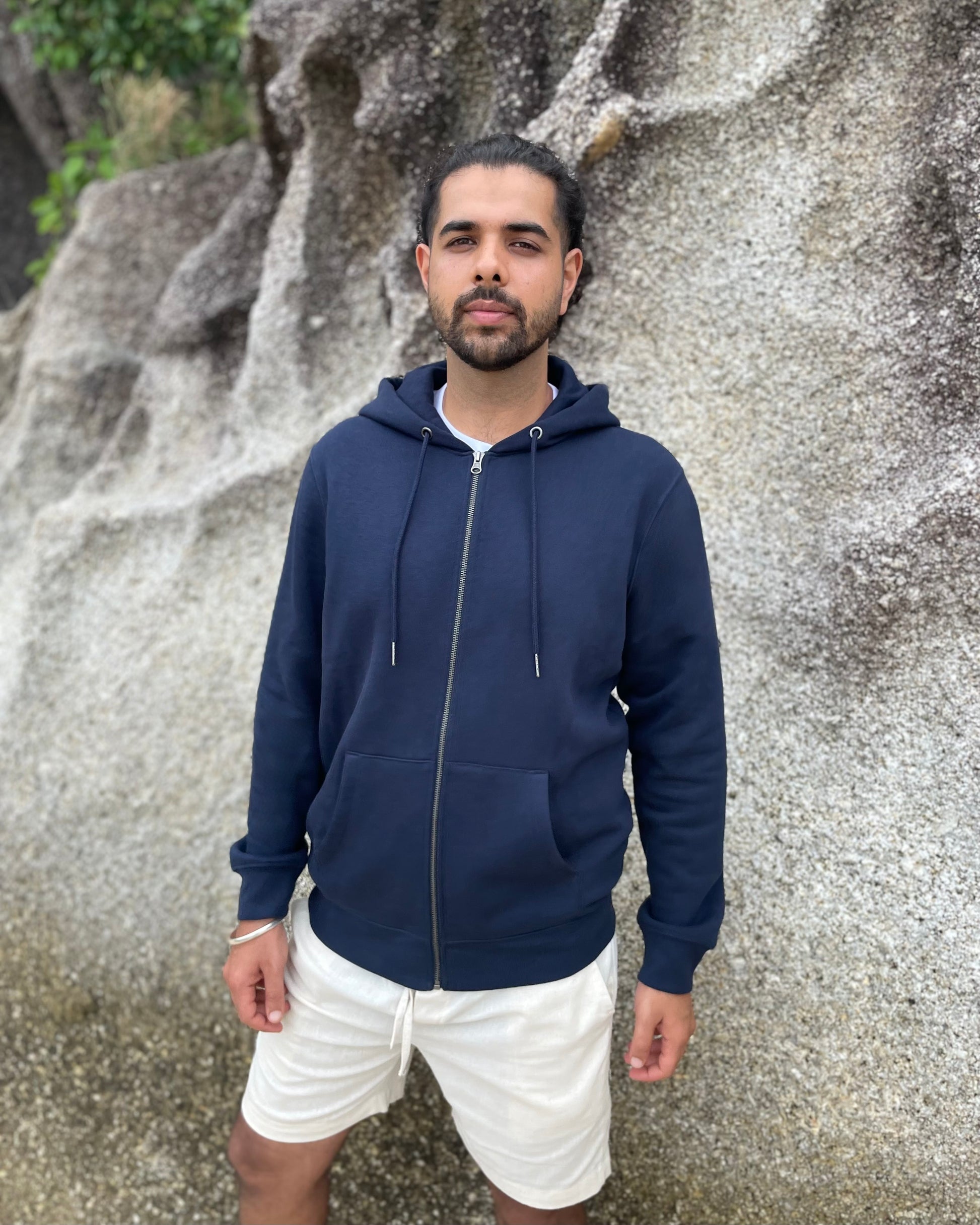 Man wearing a navy blue hoodie and white shorts standing against a rocky background