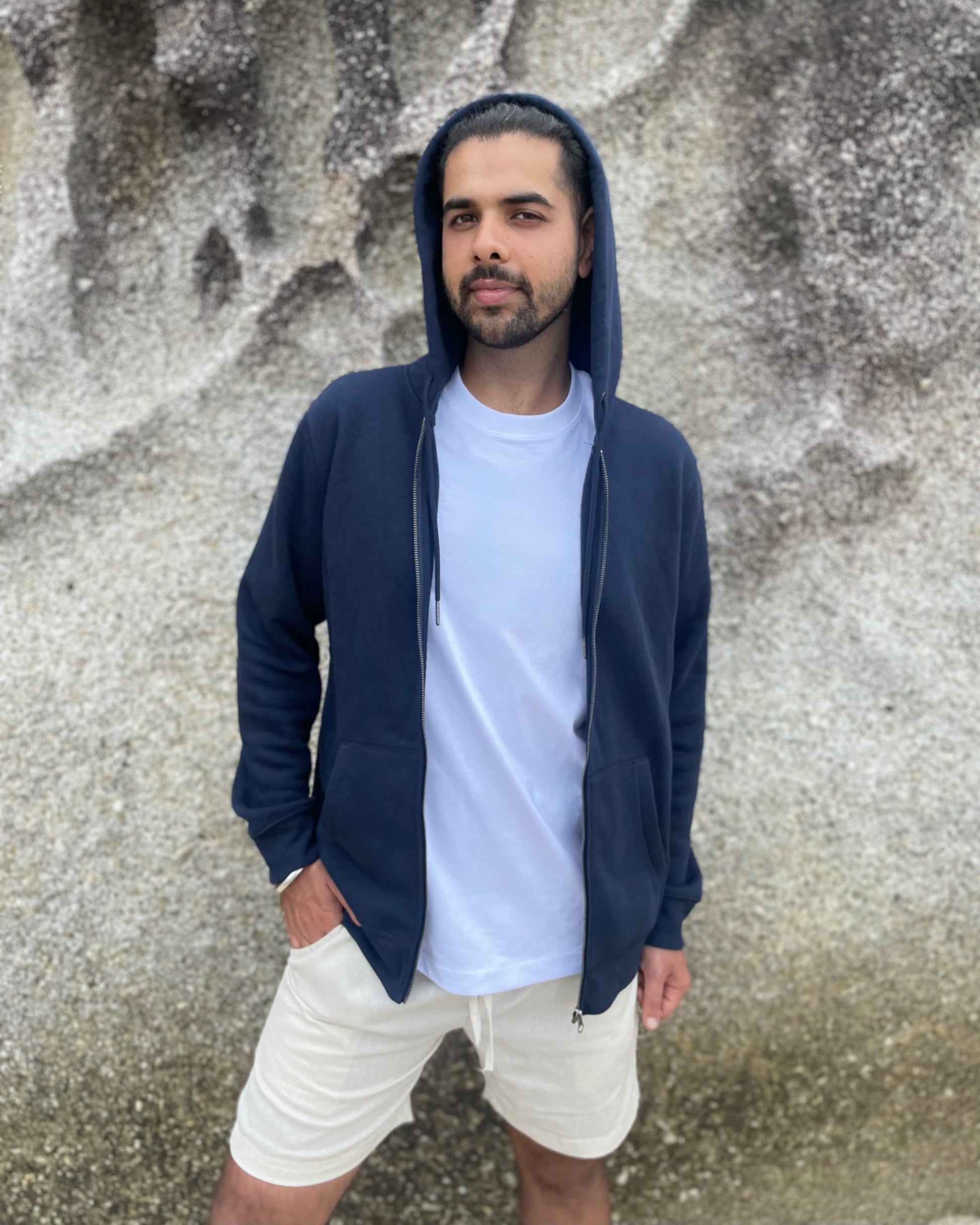 Man wearing a navy blue hoodie, white t-shirt, and beige shorts against a textured wall.
