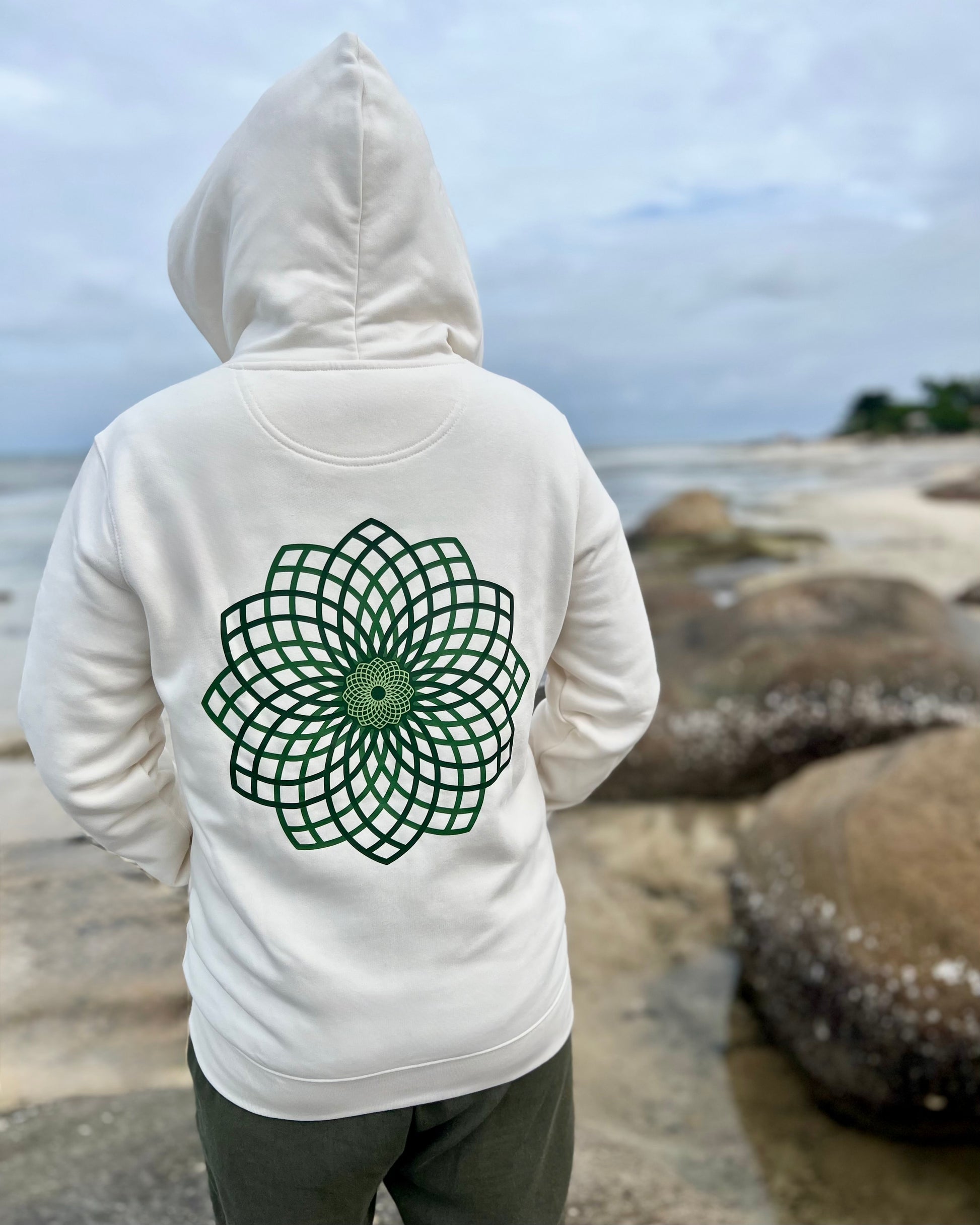 Person wearing a white hoodie with a green geometric design on the back, standing on a rocky beach.