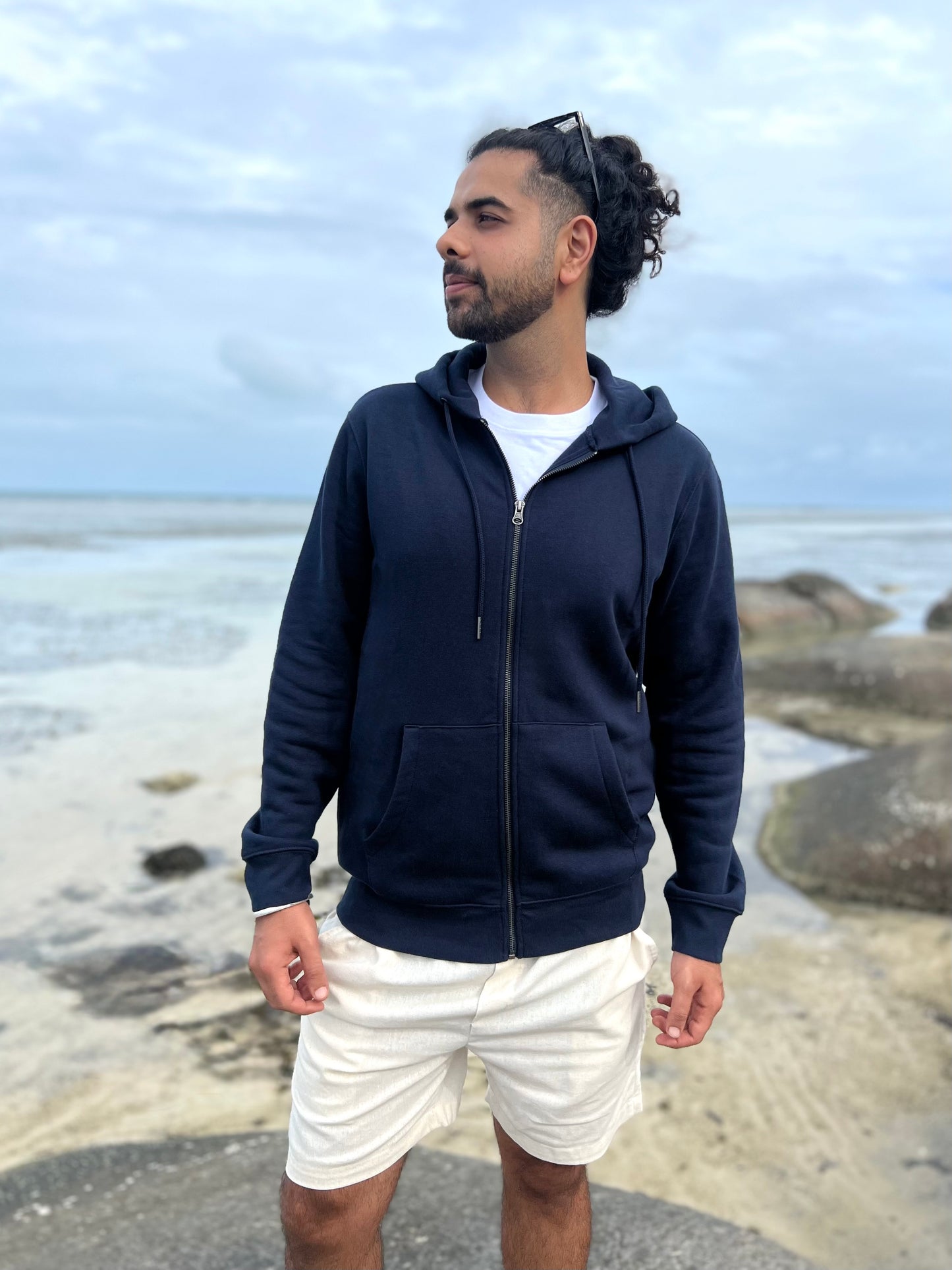 Man wearing a navy blue hoodie and white shorts standing on a rocky beach.