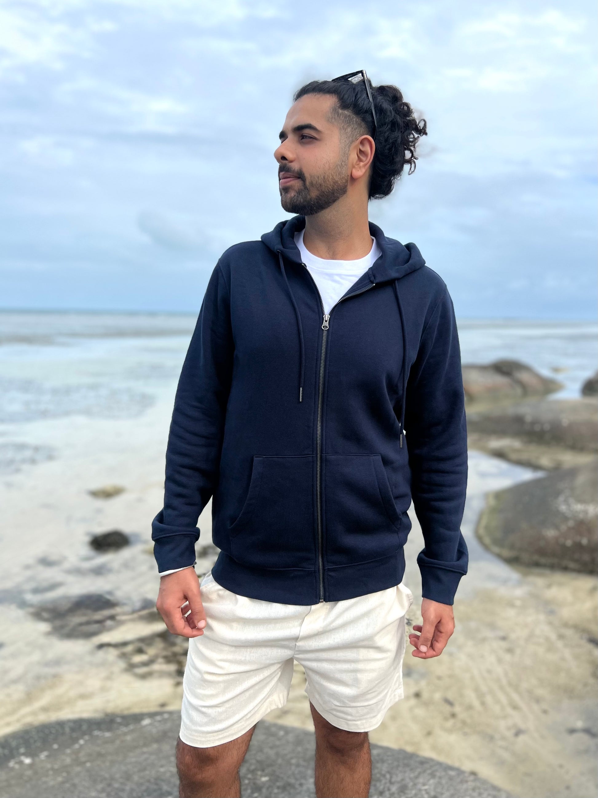 Man wearing a navy blue hoodie and white shorts standing on a rocky beach.