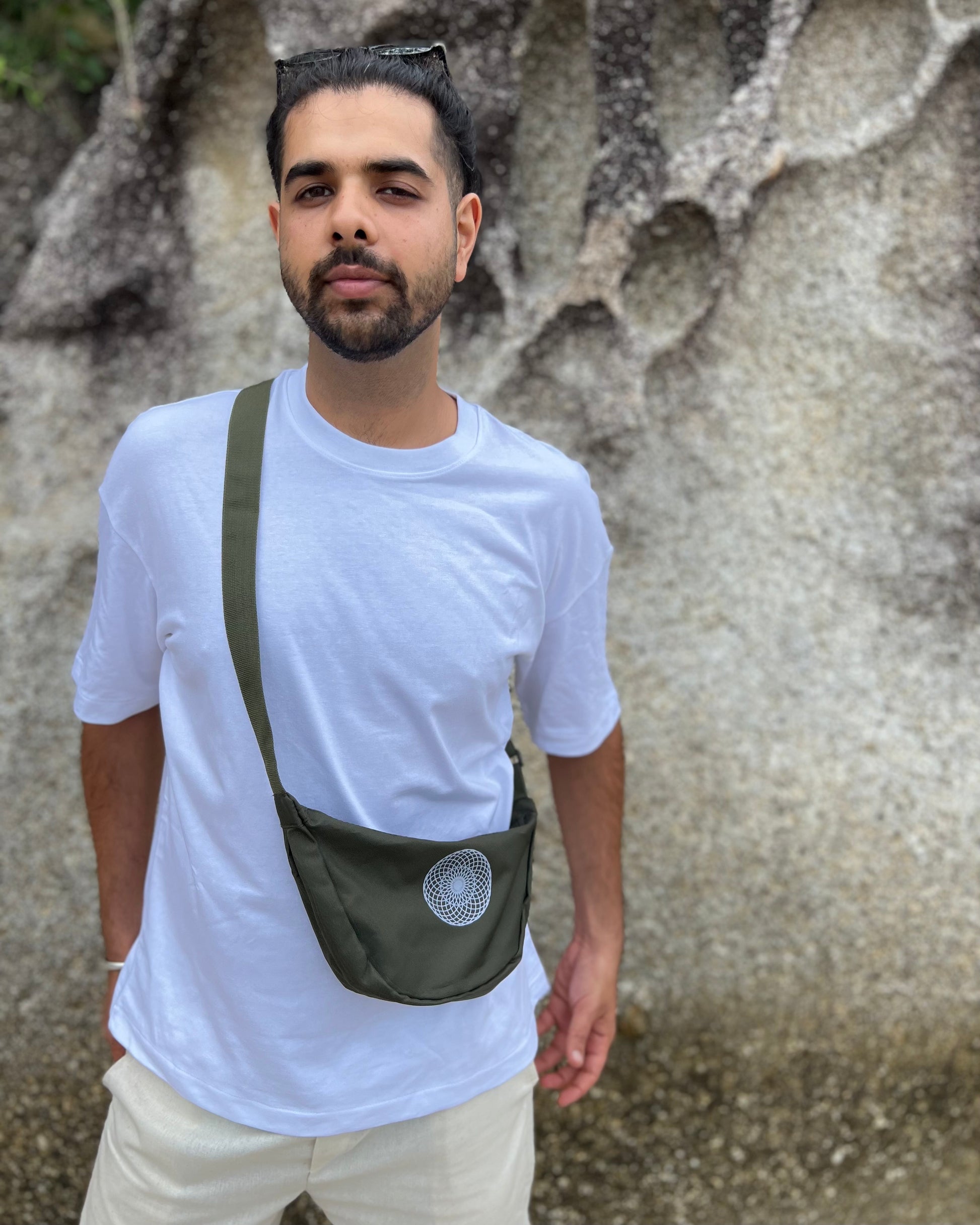 Man wearing a white t-shirt and carrying a green crossbody bag against a textured wall.